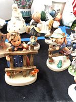 Photo showing multiple Hummel figurines featuring children and a wooden fence, along with other porcelain figurines in the background.