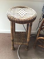 Woven rattan plant stand with circular top and lower woven shelf shown from side angle against white wall and carpeted floor.