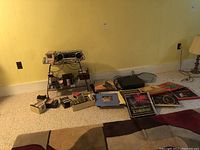Wide view of all items including boombox on stand, cassette tapes on the floor, vinyl records spread out on carpet, and electronics stacked at side.