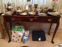 Full view of wooden side table holding various silver-plated decorative items. Three front drawers visible, only center drawer operable. Scuffs and scratches visible on table surface.