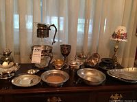 Full view of multiple silverplate and pewter items on tabletop including bowls, teapot, teabag strainer, sugar bowl, Wilton pewter plate, pewter tray, trivet, and table lamp with floral shade.