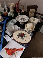 Table with various ceramic and porcelain figurines, decorative plates, and bowls visible, including Belleek vase, Goebel and Lenox pieces with holiday and animal motifs.
