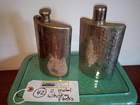 Two vintage metal whiskey flasks on a green surface with a handwritten auction tag reading "2 metal whiskey flasks."