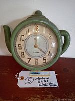 Full front view of the antique Victor teapot wall clock showing its green teapot-shaped metal casing, clock face with numbers and hands, and overall condition.