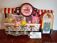 Full view of vintage wall clock depicting 1950's soda fountain with two seated figurines, ice cream counter, menu board, Coca Cola branding, and jukebox on right.
