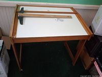 Drafting table desk viewed from an angle showing the wooden frame and white drawing surface with T-square on top.