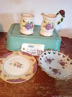 Photo showing creamer and sugar set atop a green box with two floral fine china plates in front