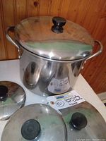 Large stainless steel cooking pot with lid and three additional lids displayed on a white surface against wooden background.