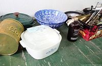 Wide view showing several items including Vintage Blue Cornflower Corning Ware casserole with glass lid, large blue floral bowl, various cooking utensils in tin, metal and ceramic pans and canisters on a green wooden surface.