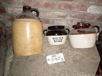 Three pottery items side by side: large tan jug with cork, white and black Boston Baked Beans pot, and brown-tan ear handle pot with lid