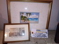 Wide view of both framed artworks stacked against a surface showing overall frames and mat condition including discoloration on the mat of the watercolor piece.