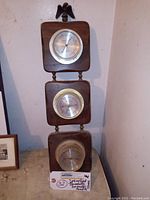 Full view of the wall hanging weather gauge showing three wood frames with instruments and eagle finial on top.