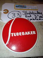 Close-up image of round red and white Studebaker car emblem showing the brand name and white vertical stripe, with tag indicating lot number and description.
