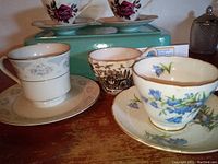 Photo showing four different tea cups and three matching saucers, including a white cup with blue floral design, a white cup with scenic pattern, and two cups with a thin brown rim.
