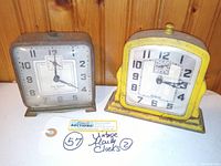 Front view showing two square vintage wind-up alarm clocks, one Gilbert in silver and one Times Square in yellow with noticeable crack