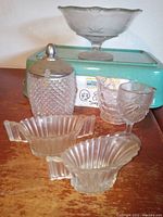 Vintage glass pieces on wooden table including lidded sugar bowl, creamers, compote dish, and two glasses