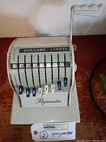 Front view of Paymaster check writer showing dollar and cent dials with currency tags and lever.