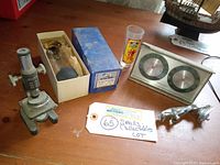 Five vintage collectibles on wood surface including microscope, boxed projector lamp, tequila glass, Springfield gauge, and Jaguar hood ornament.