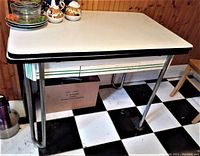 Shows the vintage white enamel kitchen table with chrome legs and black trim on top, kitchen setting with black and white floor.