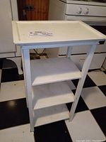 Angled view of the full white wooden shelf stand showing four shelves and its structure on a checkered floor.
