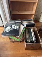 Photo showing multiple stacks of vinyl albums and boxed sets in cardboard boxes and on a small table, with varying album covers visible