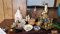 Full view of various Asian and religious figurines, brass weights, Herend Hungarian ashtray, glass knobs, and other collectibles arranged on shelf.