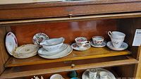 Overall display of assorted china including plates, teacups, saucers, and gravy boats arranged on a wooden shelf.