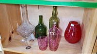 Shelf containing a clear crystal decanter with stopper, two green glass bottles, two pink cut-glass lidded containers, and a red Asian style art glass vase.