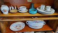Shelf showing soup tureen, platter, figurines, small plates, coffee cup and saucer, and ceramic bowl.