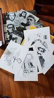 View of assortment of black and white photographic poster cards featuring 1950s-60s celebrities, laid out on a wooden surface alongside caricature prints.
