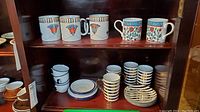 Photo showing 4 white mugs with Egyptian-style floral designs and 4 white mugs with blue interiors and floral botanical designs on the left side. Below them are enamelware cups and plates in black and white stripes.