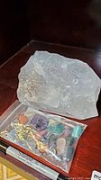 Photo showing large raw quartz stone next to a sealed plastic bag containing various small tumbled stones placed on a shelf with a Sharpie marker for scale.