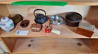 Photo of shelf with 9 Asian themed trinket boxes and home decor items including a ceramic ginger jar, cast iron teapot, wooden and metal bowls and boxes.