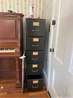 Full frontal view of tall metal file cabinet with five drawers, one drawer open at bottom showing interior storage space and metal construction, scratches and vintage wear visible on surface.