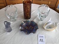 Grouping of two clear glass baskets, amber-brown vase, M.D. paperweight dish, small lavender pitcher, and white swan figurine on table