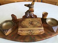 Photo showing a hand-painted wooden tray with floral designs holding two round brass ashtrays, a wooden box with floral painting, a small wooden cup, and a wooden animal figure with a pipe.