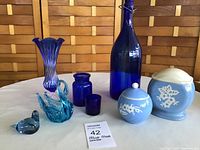 Photo of combination blue glass and pottery items on table. Includes two small bird figurines, multiple blue glass vases and jars, and round blue pottery jars with white floral designs.