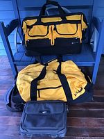 Two large yellow LLBean duffle bags with black trim on a chair, one showing exterior pockets, along with one black rectangular bag below them