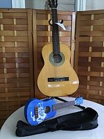 Full view of Castillo acoustic guitar on Ultra stand, blue ukulele on table with black soft case laid flat