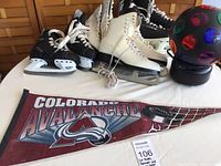 Photo shows four pairs of ice skates (two white women's pairs, two black pairs including one child-sized), Colorado Avalanche hockey pennant laid flat, and a black plastic disco light with colored lenses on a table.