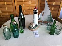 Photo showing entire lot on round table, including Lefton lighthouse figurine, metallic silver sailboat sculpture, and assortment of green glass bottles and candleholders.