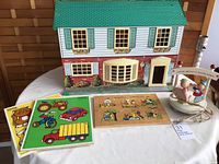 Photo showing the front of the vintage metal dollhouse with a green roof and red brick lower facade alongside the lamp and three wooden puzzles arranged on a table.