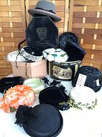 Full view of hats arranged with two hat boxes, one Disney branded and one Caravaglio branded, showing variety of decorative vintage hats in black, white, gray, and floral colors