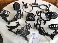 Group photo of various cast iron black metal household fixtures including lamp holders, napkin holder, towel ring, and trivet arranged on a white tablecloth.
