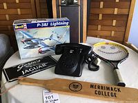 Photo showing black rotary dial telephone, Merrimack College wooden paddle, tennis racquet, Revell P-38J Lightning model kit box, Goslin's Jaguar license plate on table with background clipboard and waste paper can