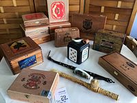 Photo showing multiple wooden cigar boxes, one tin ceramic box with playing card designs, and two decorative pipes in front of the boxes