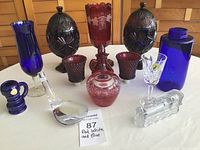 Full view of assorted red, white (clear), and blue glassware items displayed on table including vases, goblets, bottles, jars, and decorative pieces.