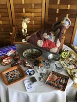 Overview of the lot displaying vintage quilt, doll bed, pottery birds, a folk art bowl, souvenir plates, a happy face clock, and various small collectibles arranged on a table
