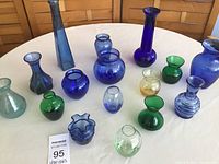 Top-down view of 16 assorted colorful vintage glass vases on round white table, showing variety of shapes, colors, and sizes, tallest about 9 inches.