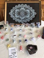 Lot overview showing Wade tea figurines in clear plastic bags arranged on a surface. Behind figurines is a wooden tray with lace fabric background, and additional small boxes and items on sides.